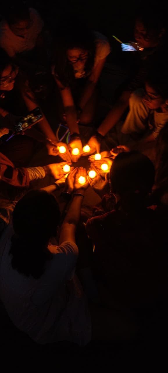 Hands cupping small glowing candles arranged in a circle on the grass, photographed from above in the dark.