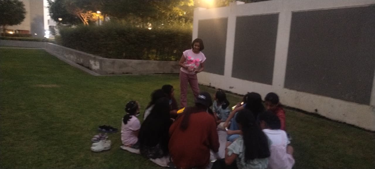 Anagha standing in front of friends seated on a mat in our community park at dusk, holding glow stick bracelets to welcome everyone.